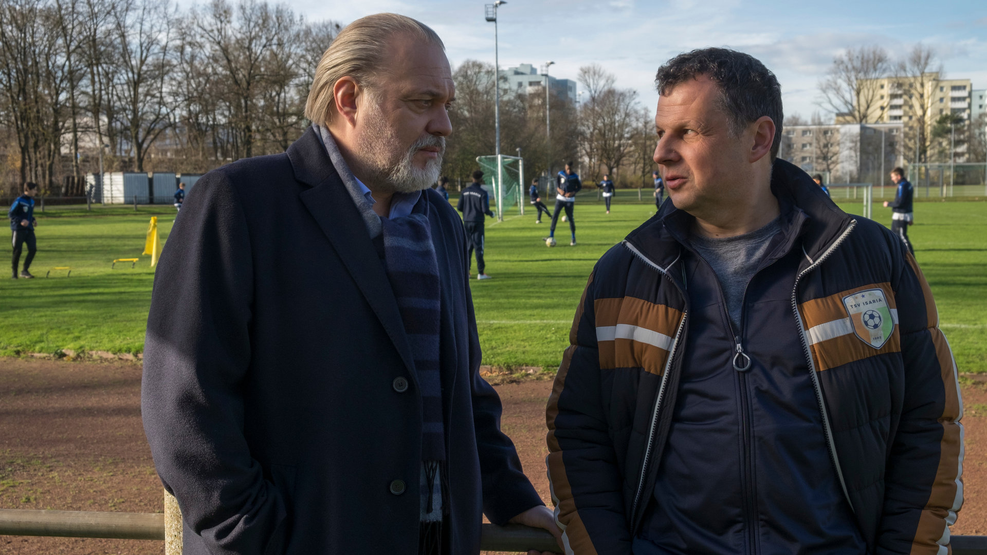 "Der Alte - Aufstiegskampf": Hauptkommissar Richard Voss (Jan-Gregor Kremp) spricht mit Dieter Heuring (Gerhard Wittmann). Die beiden stehen an der Bande mit dem Rücken zu einem Fußballplatz, auf dem Nachwuchsfußballer trainieren.