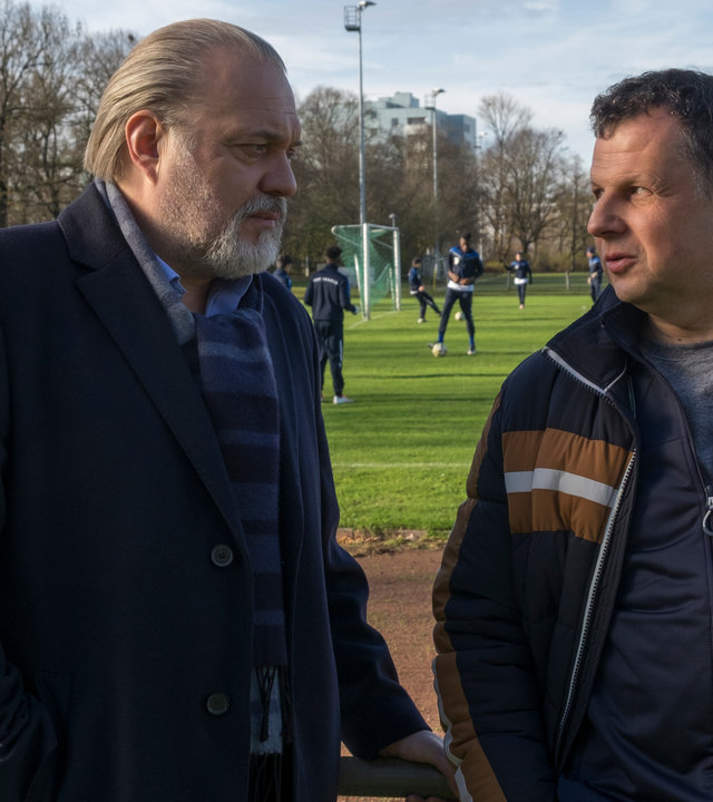 "Der Alte - Aufstiegskampf": Hauptkommissar Richard Voss (Jan-Gregor Kremp) spricht mit Dieter Heuring (Gerhard Wittmann). Die beiden stehen an der Bande mit dem Rücken zu einem Fußballplatz, auf dem Nachwuchsfußballer trainieren.