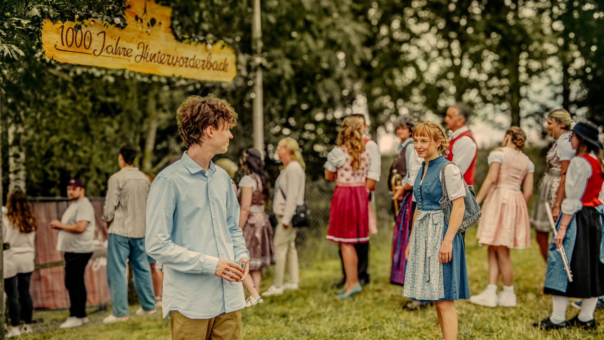 "Tschappel - 1000 Jahre Hintervorderbach": Carlo (Jeremias Meyer) steht in Hemd und Hose bekleidet auf einer Festwiese und blickt zu Pia (Mina-Giselle Rüffer), die ein Dirndl trägt und eine Tasche über der Schulter trägt. Im Hintergrund sind festlich gekleidete Menschen (Komparsen), einige in Tracht, über ihnen hängt ein Holzschild mit der Aufschrift "1000 Jahre Hintervorderbach".