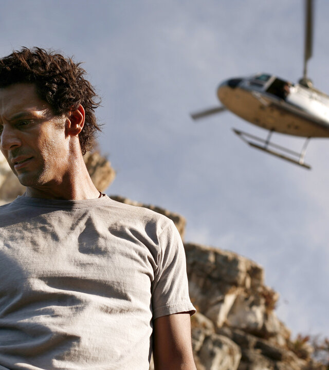 "Largo Winch - Tödliches Erbe": Largo Winch (Tomer Sisley) steht in einer felsigen, kargen Landschaft. Über ihm fliegt ein Hubschrauber.