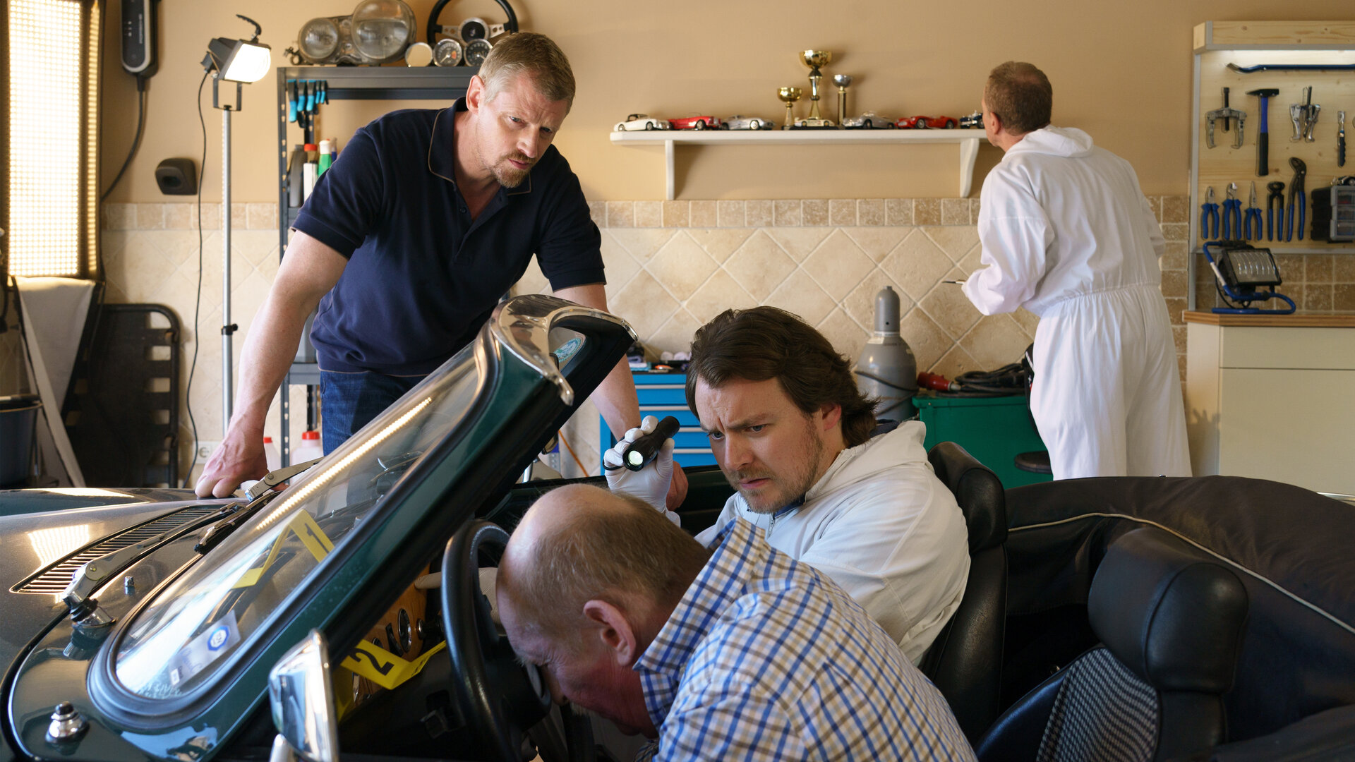 "SOKO Wien - Vergeltung": In einer Garage: Major Max Herzog (Martin Gruber) stützt sich mit beiden Armen am Tatfahrzeug ab und beobachtet Spurensicherer Julius Rubatsch (Max Fischnaller) bei der Untersuchung der Leiche von Werner Novotny (Franz Bohac). Werner Novotny sitzt auf dem Fahrersitz und sein Kopf lehnt am Lenkrad. Julius Rubatsch hält in der linken Hand ein kleine schwarze Taschenlampe und leuchtet damit den Kopf von Werner Novotny an. Im Hintergrund untersucht ein weiterer KTU-Mitarbeiter (Komparse) ein Regal mit kleinen Sammelautos.