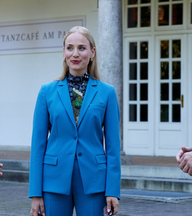 "Die Rosenheim-Cops - Ein riesiges Problem": Die Kommissare Kaya (Baran Hêvî) und Hansen (Igor Jeftić) stehen vor der weißen Eingangstür eines Gebäudes und sprechen mit Sarah Niedermeier (Louisa von Spies). Neben der Eingangstür ist ein Schild mit der Aufschrift "Tanzcafé am Park" angebracht.