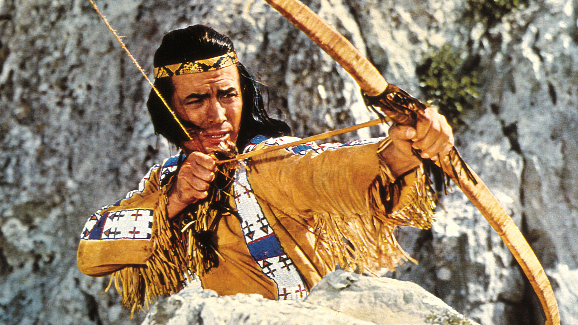 "Winnetou und sein Freund Old Firehand": Großaufnahme von Winnetou (Pierre Brice) mit einem gespannten Bogen vor einer Felslandschaft.