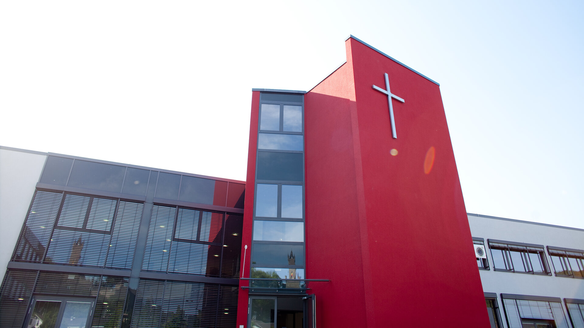 "Evangelischer Gottesdienst - Von Gott beschenkt. Von Gott bewegt.": Außenansicht des Gemeindezentrums der Freien Evangelischen Gemeinden Dillenburg: Bei einem modern gestalteten, länglichen Gebäude befindet sich ein roter stilisierter Kirchenturm mit einem großen Kreuz an der Vorderfront in der Mitte.