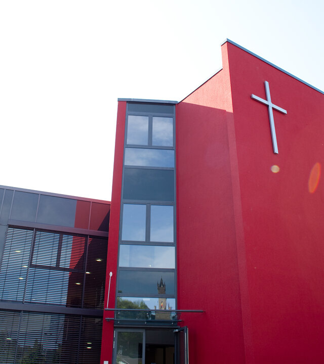 "Evangelischer Gottesdienst - Von Gott beschenkt. Von Gott bewegt.": Außenansicht des Gemeindezentrums der Freien Evangelischen Gemeinden Dillenburg: Bei einem modern gestalteten, länglichen Gebäude befindet sich ein roter stilisierter Kirchenturm mit einem großen Kreuz an der Vorderfront in der Mitte.