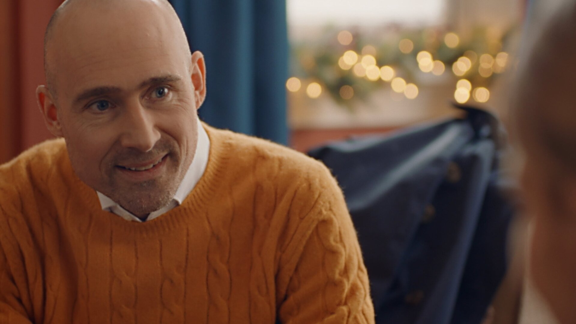 "Single Bells - 12 Dates till Christmas: Hendrik": Hendrik (Geerard Van de Walle) in Nahaufnahme. Er hat eine Glatze und trägt einen senfgelben Wollpulli über einem weißen Hemd, dabei blickt er freundlich lächelnd zur Seite.