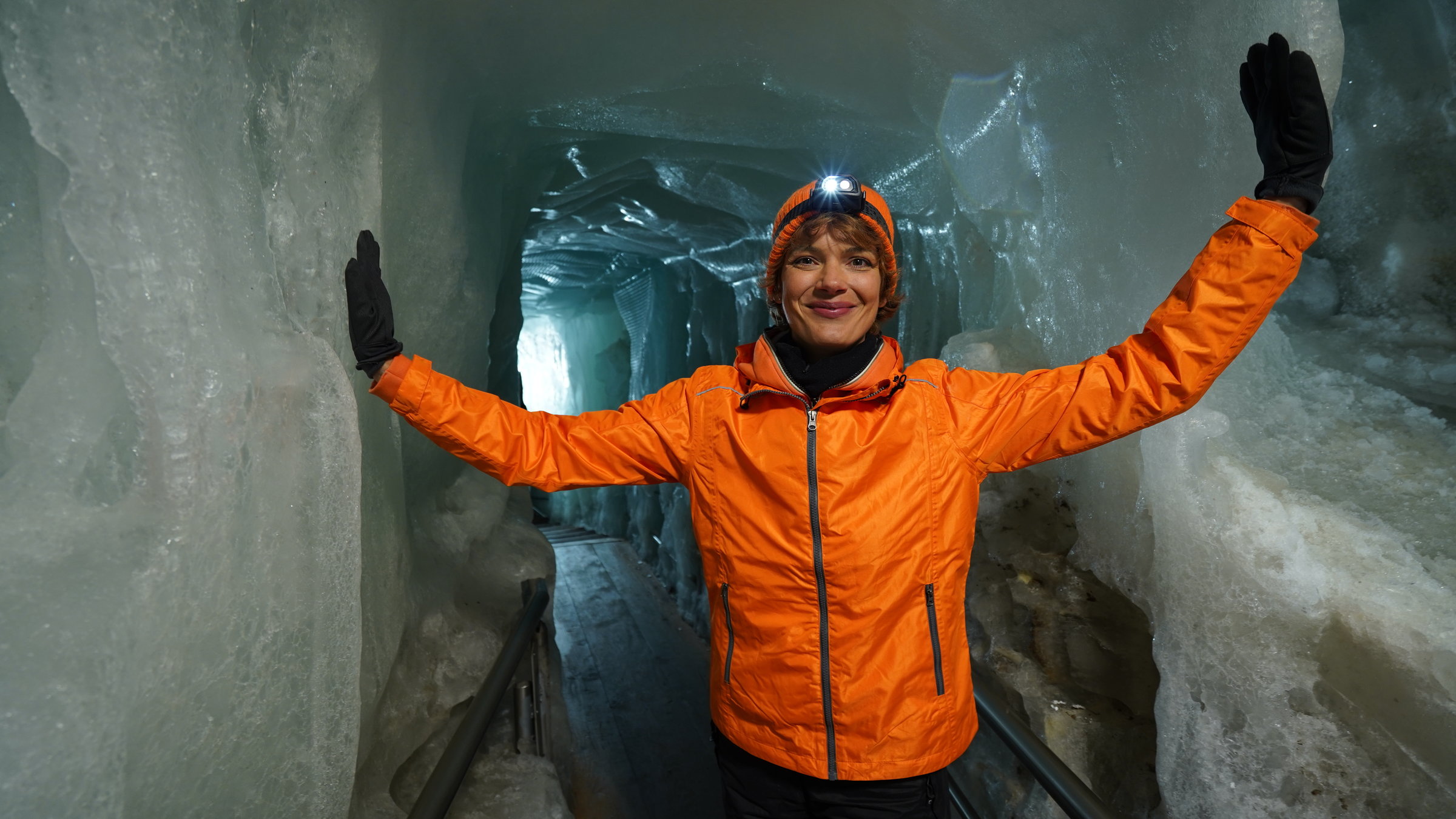 "Terra Xpress: Faszination Alpen - Zerbrechliches Paradies": Moderatorin Lena Ganschow steht zwischen zwei Eiswänden und hat jeweils eine Hand an einer Eiswand. Sie trägt eine orangefarbene Jacke und einen Helm.