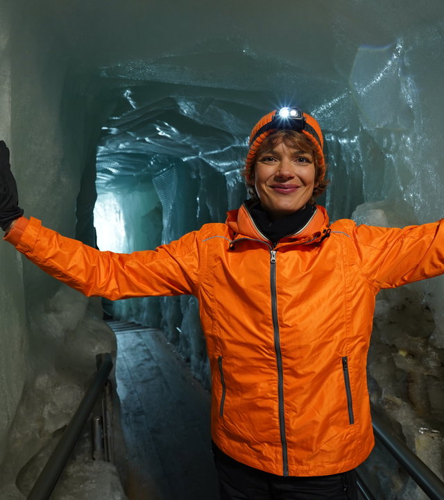 "Terra Xpress: Faszination Alpen - Zerbrechliches Paradies": Moderatorin Lena Ganschow steht zwischen zwei Eiswänden und hat jeweils eine Hand an einer Eiswand. Sie trägt eine orangefarbene Jacke und einen Helm.