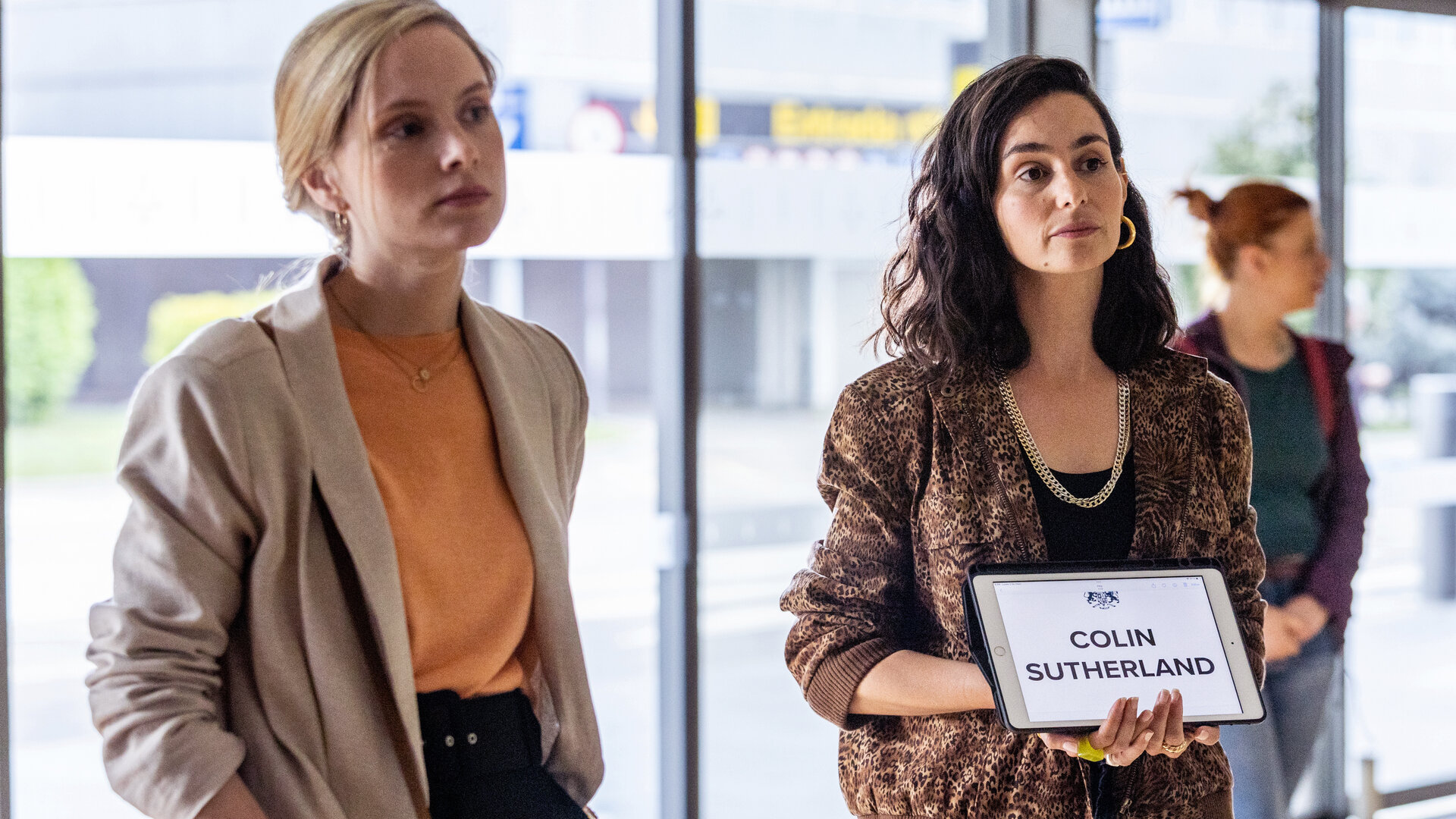 "The Diplomat - Tod in Barcelona": Laura Simmonds (Sophie Rundle) und Alba Ortiz (Serena Manteghi) stehen nebeneinander im Wartebereich des Flughafens. Alba Ortiz hält ein Schild mit der Aufschrift "COLIN SUTHERLAND" sichtbar in der Hand.