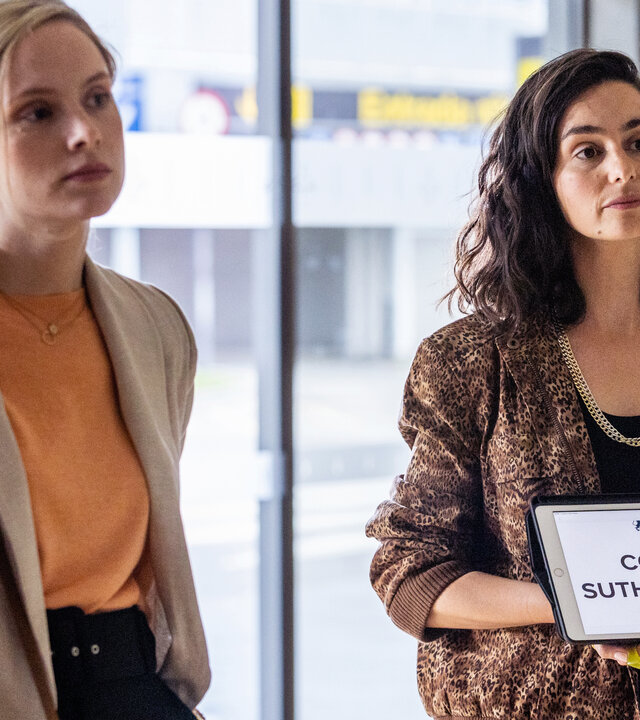"The Diplomat - Tod in Barcelona": Laura Simmonds (Sophie Rundle) und Alba Ortiz (Serena Manteghi) stehen nebeneinander im Wartebereich des Flughafens. Alba Ortiz hält ein Schild mit der Aufschrift "COLIN SUTHERLAND" sichtbar in der Hand.