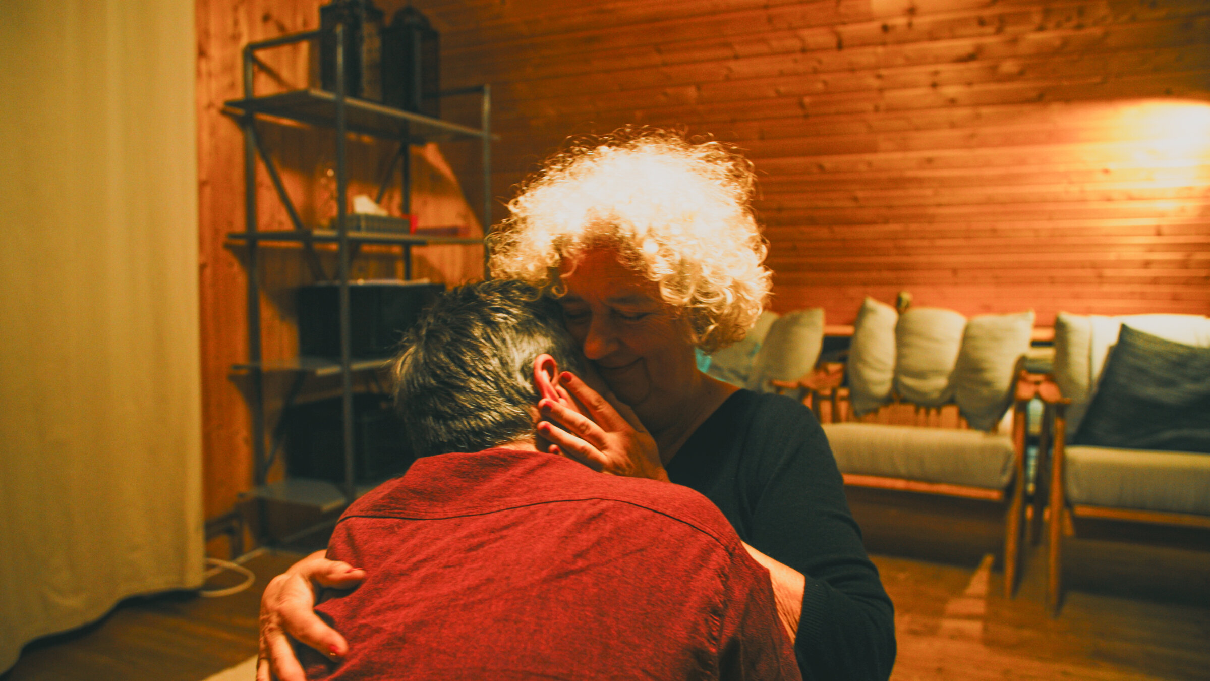 "Weiblich, Ü50 und viele Liebhaber": In einem Raum mit einer Couch im Hintergrund, sitzt Sandra auf einem Stuhl. Ein Mann kniet vor ihr, den Rücken zur Kamera gewandt. Sie hält ihn im Arm.