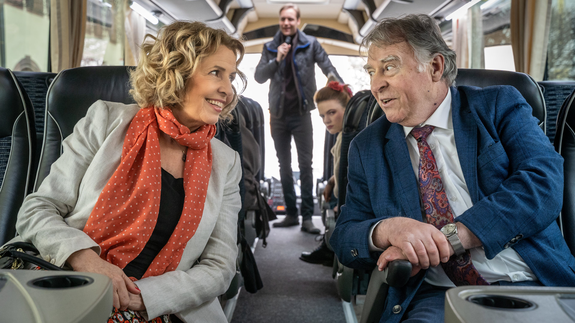 "SOKO Leipzig - Auftrag für Hajo": Maria Schneider (Michaela May) und Hajo Trautzschke (Andreas Schmidt-Schaller) sitzen in einem Reisebus nebeneinander und unterhalten sich. Thorsten Filbinger (Bernd-Christian Althoff) steht im Hintergrund des Bildes mit einem Mikrofon in der Hand. Jule Bär (Luzie Juckenburg) sitzt einige Reihen hinter Hajo und Maria und beobachtet die beiden.