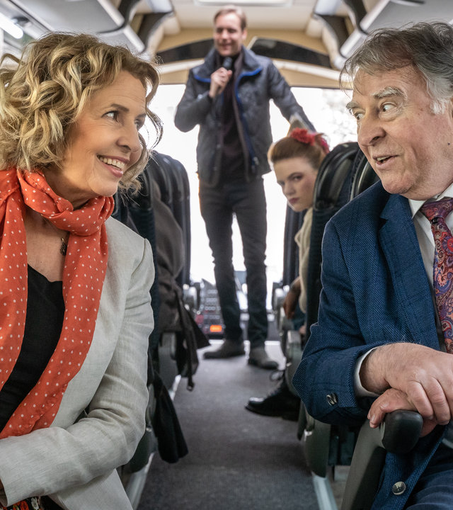 "SOKO Leipzig - Auftrag für Hajo": Maria Schneider (Michaela May) und Hajo Trautzschke (Andreas Schmidt-Schaller) sitzen in einem Reisebus nebeneinander und unterhalten sich. Thorsten Filbinger (Bernd-Christian Althoff) steht im Hintergrund des Bildes mit einem Mikrofon in der Hand. Jule Bär (Luzie Juckenburg) sitzt einige Reihen hinter Hajo und Maria und beobachtet die beiden.