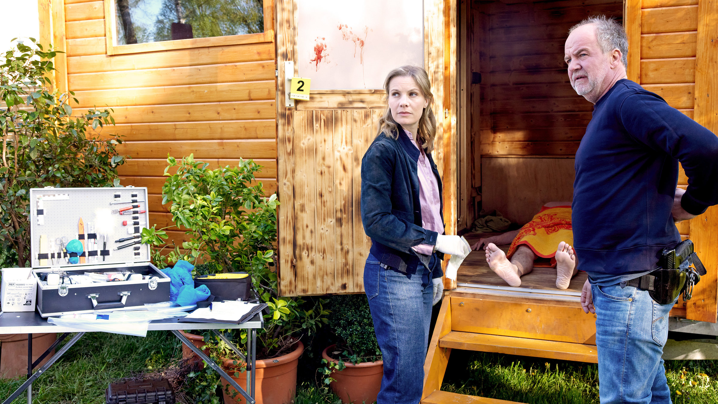 "SOKO Hamburg - Diagnose Mord": Oskar Schütz (Marek Erhardt) und Lena Testorp (Anna von Haebler) stehen sich vor einer Saune in einem Garten gegenüber und blicken ernst nach links. Sie stehen seitlich zur Kamera. Zwischen ihnen sieht man in der geöffneten Sauna die Beine und Füße eines Menschen, der auf dem Rücken in der Sauna liegt und über dem ein gelb-rotes Handtuch liegt. Links im Bild steht ein geöffneter Koffer mit diversen Utensilien auf einem Klapptisch. Um den Koffer herum liegen Unterlagen.