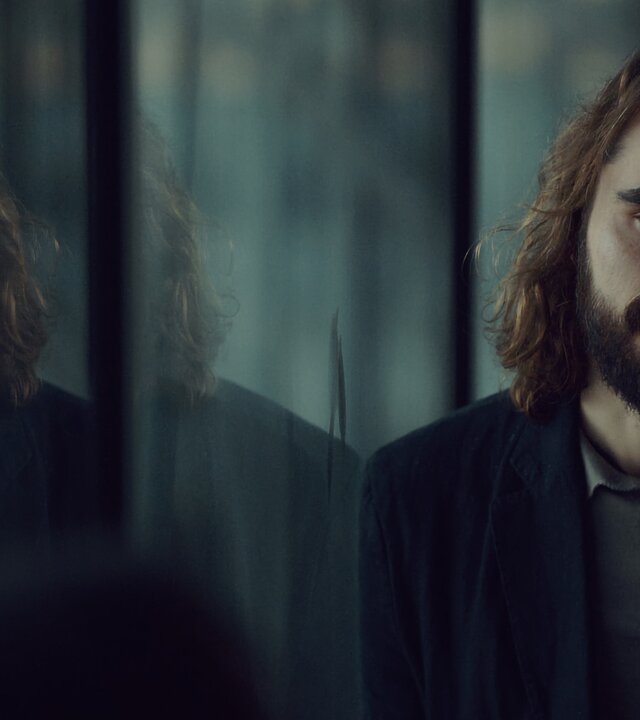 "Moresnet - The Book of Death: Der Preis der Erinnerung": Ben Schotz (Boris Van Severen) steht neben einer großen Spiegelfläche und schaut gedankenverloren nach vorne. Links von ihm ist sein Spiegelbild zu erkennen.