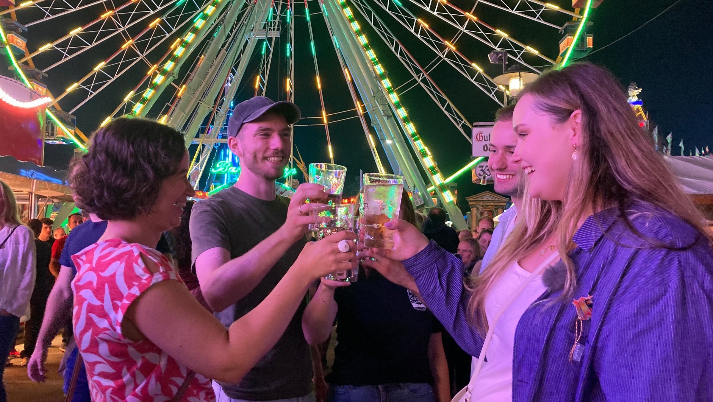 "ZDF.reportage: Wein, Wurst und Riesenrad - Das größte Weinfest der Welt": Luisa und ihre vier Freunde stehen im Kreis und stoßen mit Dubbegläsern an. Im Hintergrund stehen Menschen, das Riesenrad, Stände und ein Teil der Achterbahn.