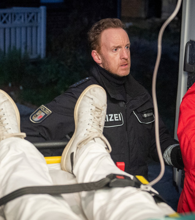 "SOKO Wismar – Tod im Rettungswagen": Timmermann (Mathias Junge) und Stine Bergendal (Sisdel Hindhede) stehen vor der geöffneten Hintertür eines Rettungswagens. Die Beine der Leiche Dr. Christian Pohls (Marco Matthes) auf der Trage sind zu sehen. Von hinten sieht man Franziska Wagner (Pinar Erincin) gefesselt.
