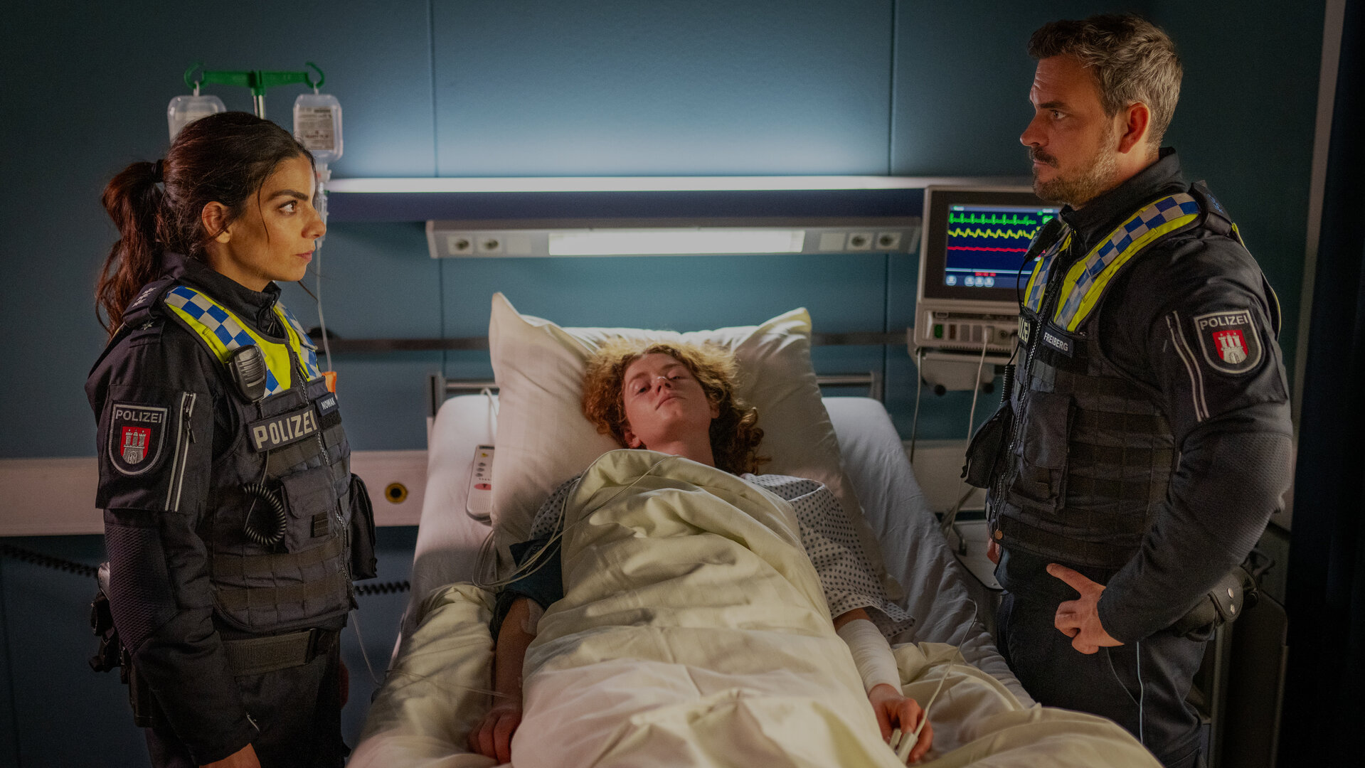 "Notruf Hafenkante - Tödliche Keime": Isa (Lilli Hollunder) und Kris (Marc Barthel) stehen links und rechts neben dem Krankenbett von Raffaela (Nadja Sabersky). Sie schauen sich an. Raffaela blickt ins Leere.