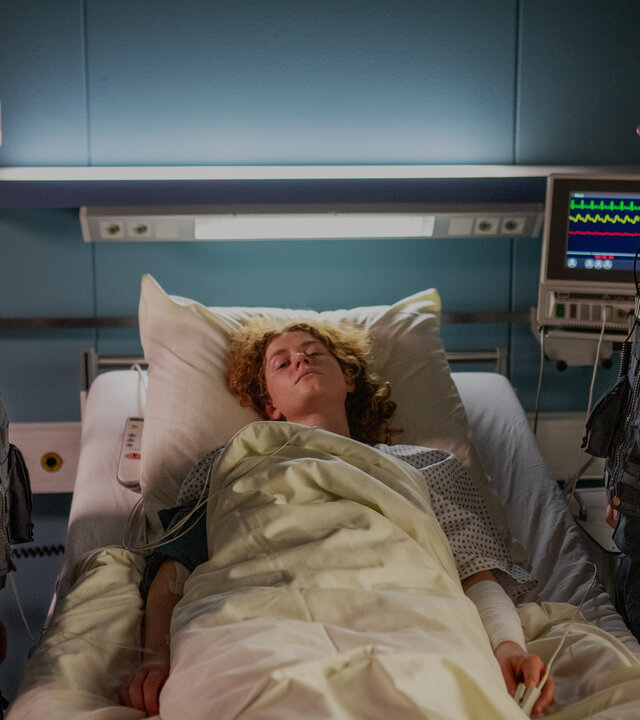 "Notruf Hafenkante - Tödliche Keime": Isa (Lilli Hollunder) und Kris (Marc Barthel) stehen links und rechts neben dem Krankenbett von Raffaela (Nadja Sabersky). Sie schauen sich an. Raffaela blickt ins Leere.