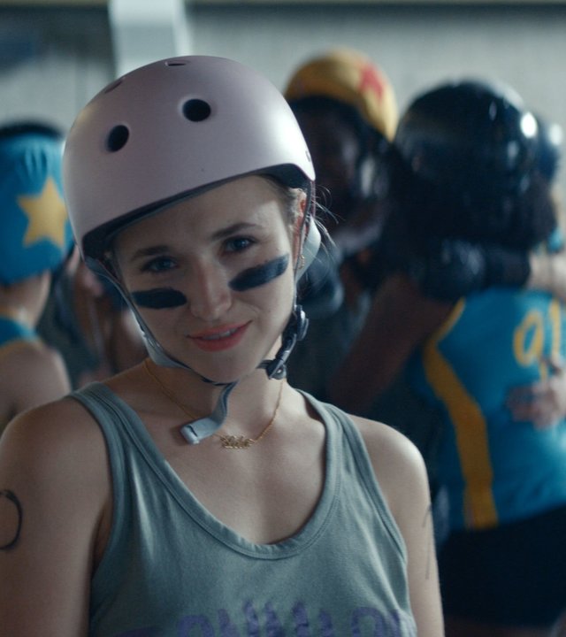 "Derby Girl - Der Pakt": Lola Bouvier (Chloé Jouannet) blickt triumphierend in die Kamera. Sie hat schwarze Streifen unter ihren Augen und trägt einen Helm. Im Hintergrund sind weitere Frauen in Sportklamotten zu sehen, die einen Kreis bilden.