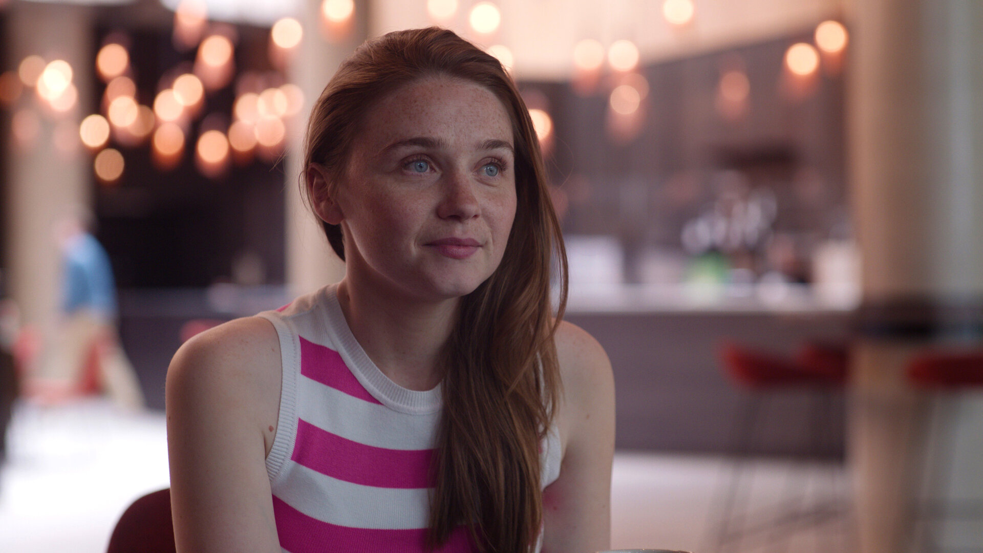 "You & Me": Emma (Jessica Barden) sitzt in einem Café und schaut lächelnd zur Seite.