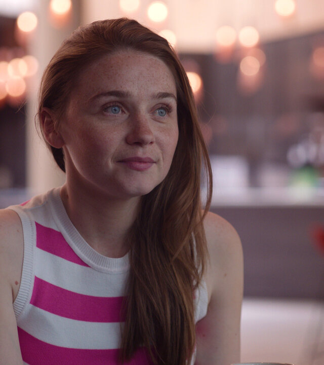"You & Me": Emma (Jessica Barden) sitzt in einem Café und schaut lächelnd zur Seite.