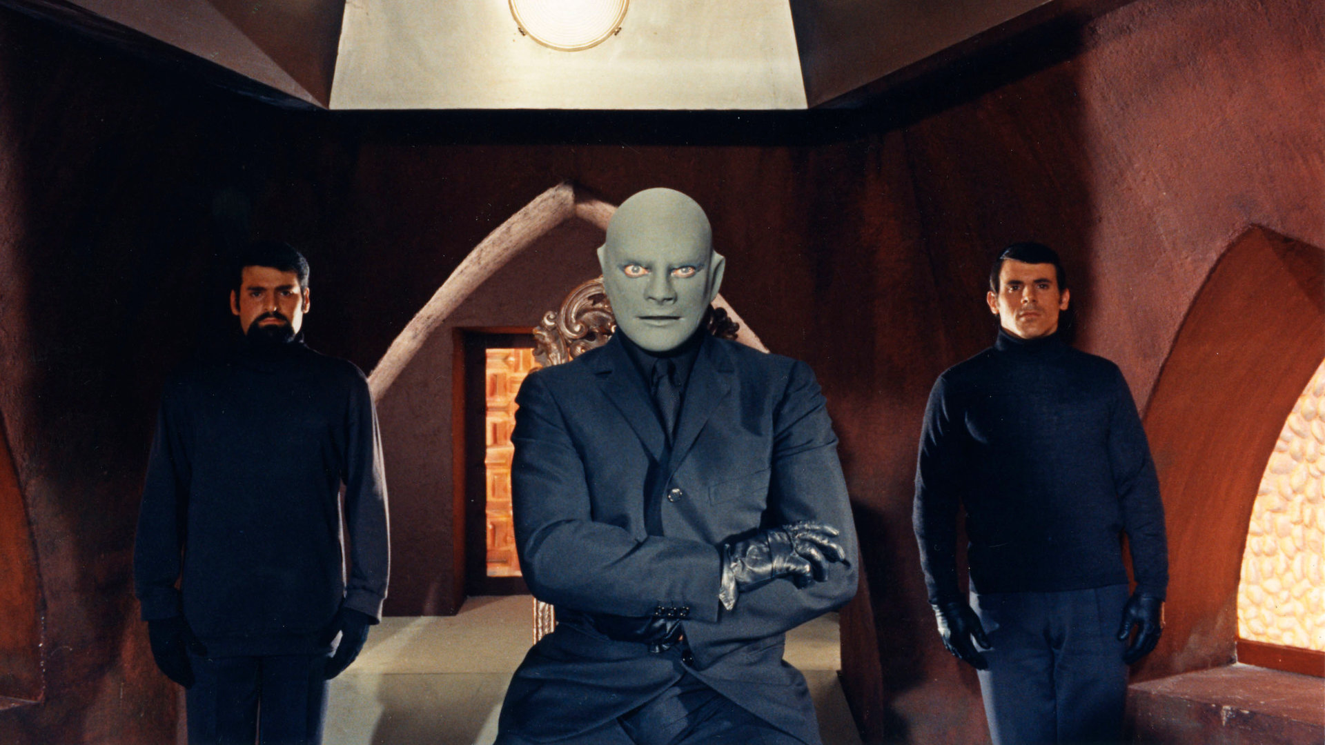 "Fantomas bedroht die Welt": Fantomas (Jean Marais) sitzt mit verschränkten Armen in einer Höhle, während Richard (Hubert de Lapparent) und der Mafiaboss (Guy Delorme) neben ihm Wache stehen.