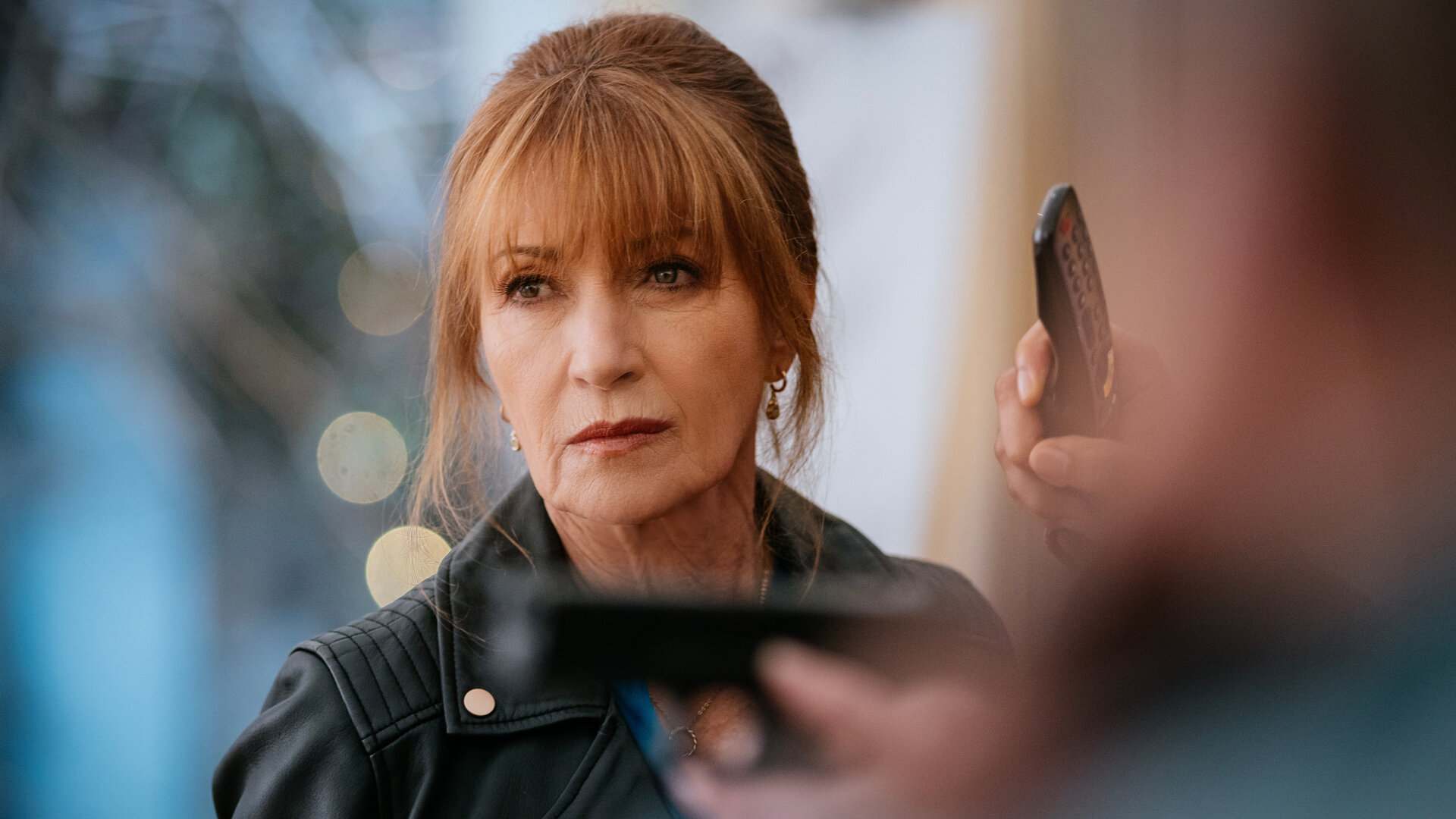 "Harry Wild – Mörderjagd in Dublin - Ein Hochzeits-Crasher aus der Hölle (2)": Harry (Jane Seymour) steht in einem Raum und hat einen ernsten Gesichtsausdruck. Vor ihr in der Unschärfe sind die Hände einer Person zu sehen, die eine Pistole auf ein Ziel neben Harry richtet.