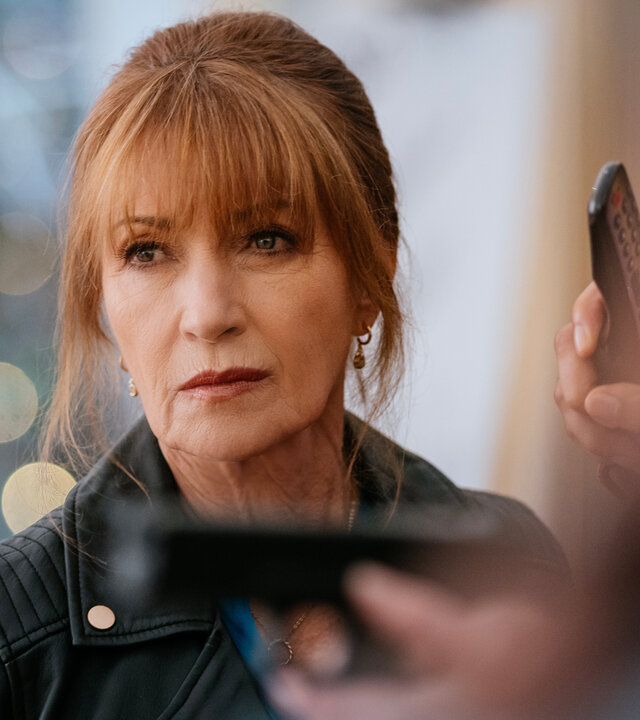 "Harry Wild – Mörderjagd in Dublin - Ein Hochzeits-Crasher aus der Hölle (2)": Harry (Jane Seymour) steht in einem Raum und hat einen ernsten Gesichtsausdruck. Vor ihr in der Unschärfe sind die Hände einer Person zu sehen, die eine Pistole auf ein Ziel neben Harry richtet.