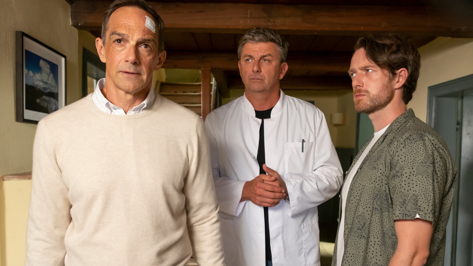 "Der Bergdoktor: Unverzeihlich (1)": In der Arztpraxis: Dr. Martin Gruber (Hans Sigl) und Elias Jung(Maximilian Klas) stehen mit fragendem Blick hinter Gero Jung (Harry Blank). Dieser trägt ein Pflaster an der Stirn und hat die Hand verbunden und wirkt entschlossen.