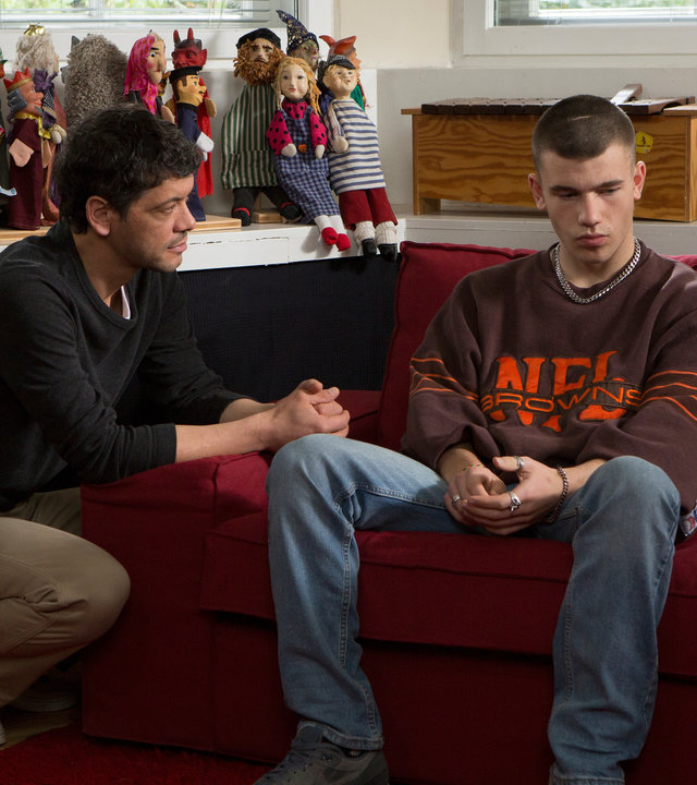 "SAFE - Episode 3": Tom (Carlo Ljubek) sitzt in der Hocke vor einem Sofa und redet mit Sam (Valentin Oppermann), der vor ihm auf einem Sofa sitzt. Im Hintergrund sind eine Fensterfront und Sideboards auf denen diverse Handpuppen und Plüschtiere stehen, sowie ein Xylophon zu sehen.