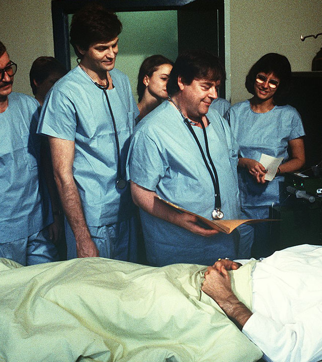 "Die Schwarzwaldklinik - Gefahr für Udo": Dr. Udo Brinkmann (Sascha Hehn) liegt im Krankenbett. Das Ärzteteam steht um das Bett herum.