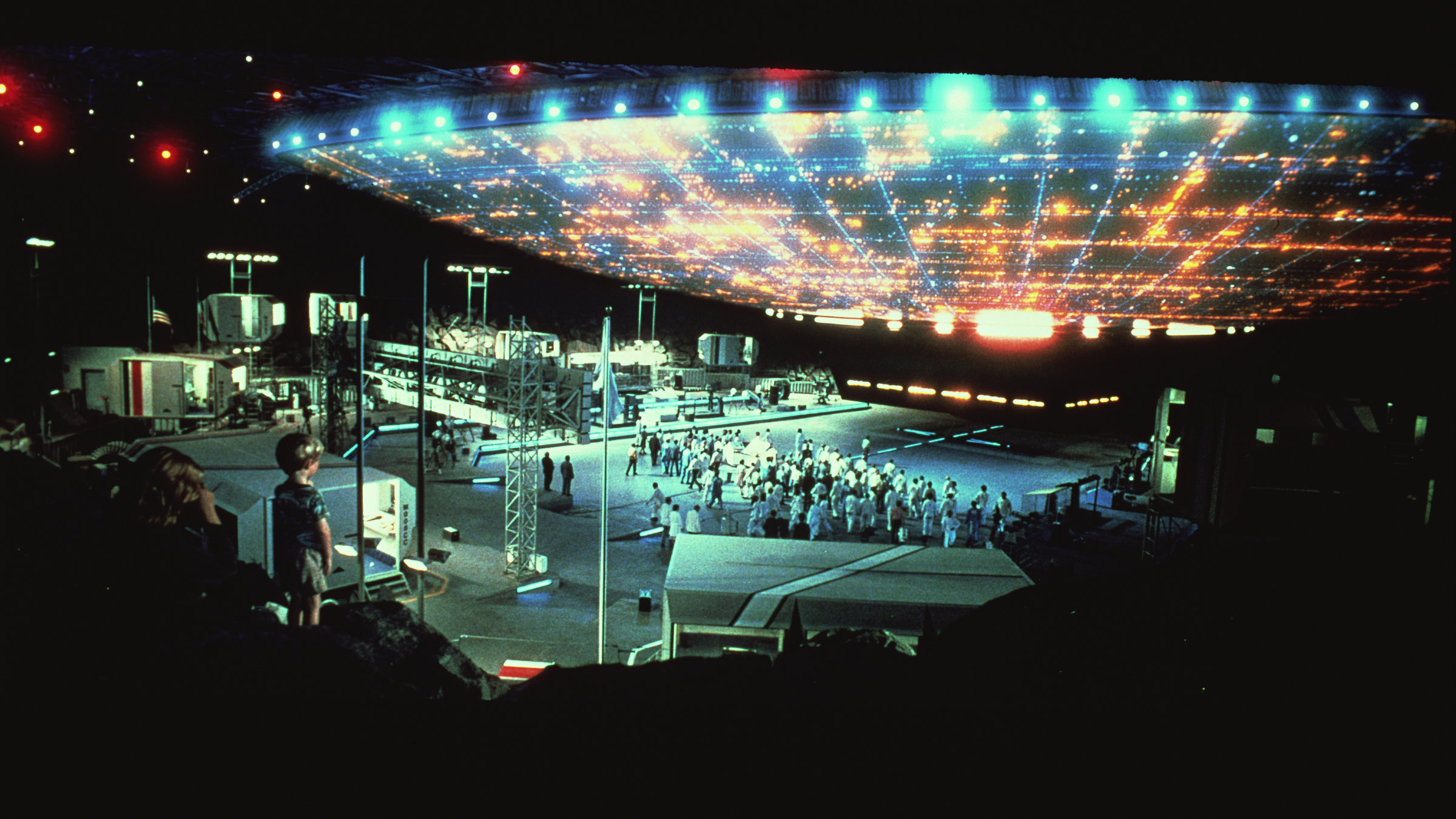 "Unheimliche Begegnung der dritten Art": Ein riesiges, hell leuchtendes Raumschiff steht knapp über dem Boden, unter ihm steht eine größere Gruppe von Wissenschaftlern in weißer Kleidung, umringt von technischen Aufbauten. Auf der linken Bildseite ist ein kleiner Junge mit seiner Mutter zu sehen, die sich das Spektakel anschauen.