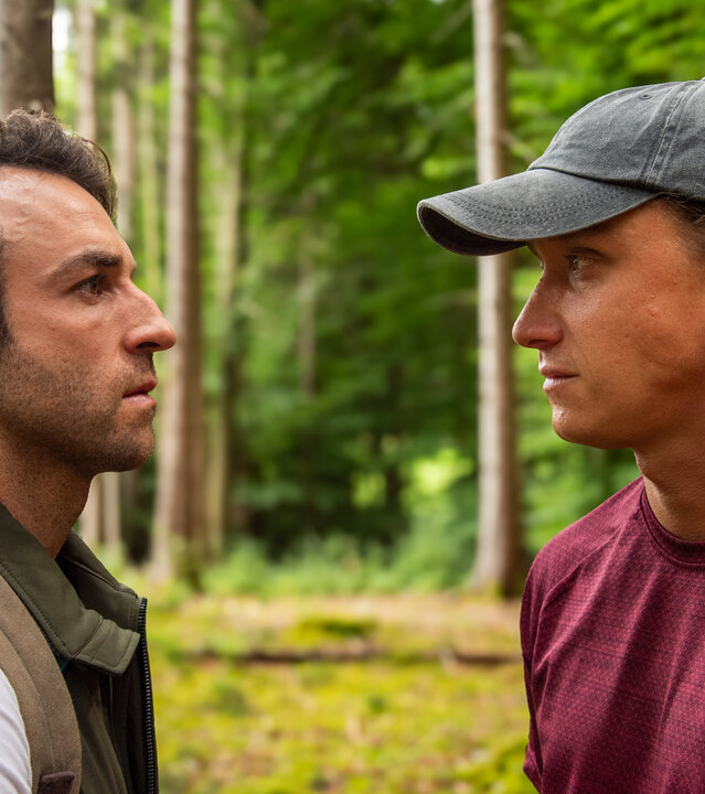 "Baumgeflüster": Yascha (Linus Schütz) und Mats (Aaron Friesz) stehen gemeinsam in einem Wald und schauen sich an.