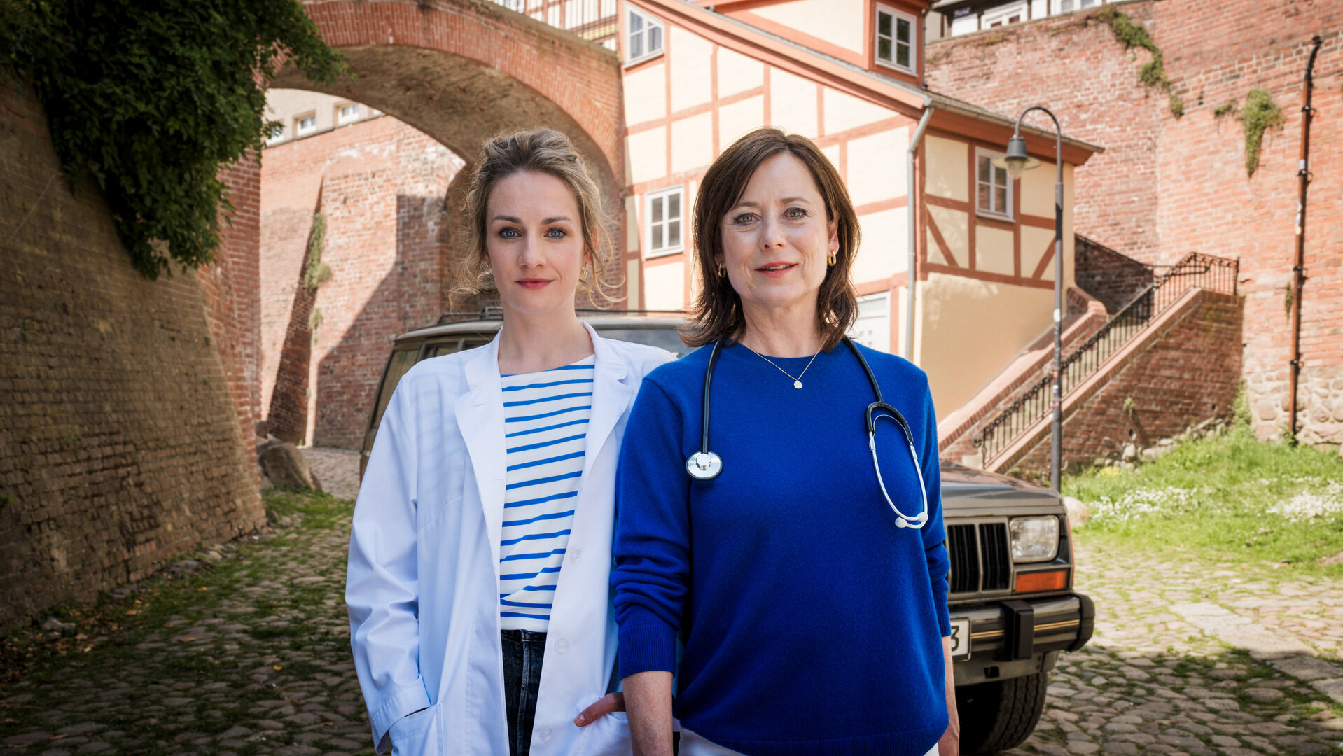 "Mit Herz und Holly: Lichtblicke": Vor einer mittelalterlichen Stadtmauer und Fachwerkhäusern stehen Dr. Holly Sass (Karoline Teska) und Dr. Katrin Herz (Inka Friedrich) und lächeln in die Kamera. Dr. Holly Sass trägt einen weißen Arztkittel. Dr. Katrin Herz hat ein Stethoskop umgehängt.