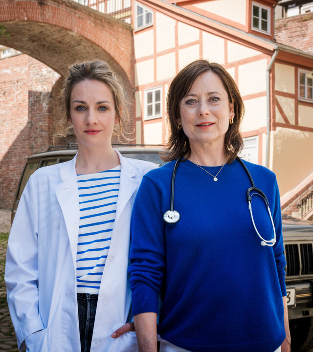 "Mit Herz und Holly: Lichtblicke": Vor einer mittelalterlichen Stadtmauer und Fachwerkhäusern stehen Dr. Holly Sass (Karoline Teska) und Dr. Katrin Herz (Inka Friedrich) und lächeln in die Kamera. Dr. Holly Sass trägt einen weißen Arztkittel. Dr. Katrin Herz hat ein Stethoskop umgehängt.