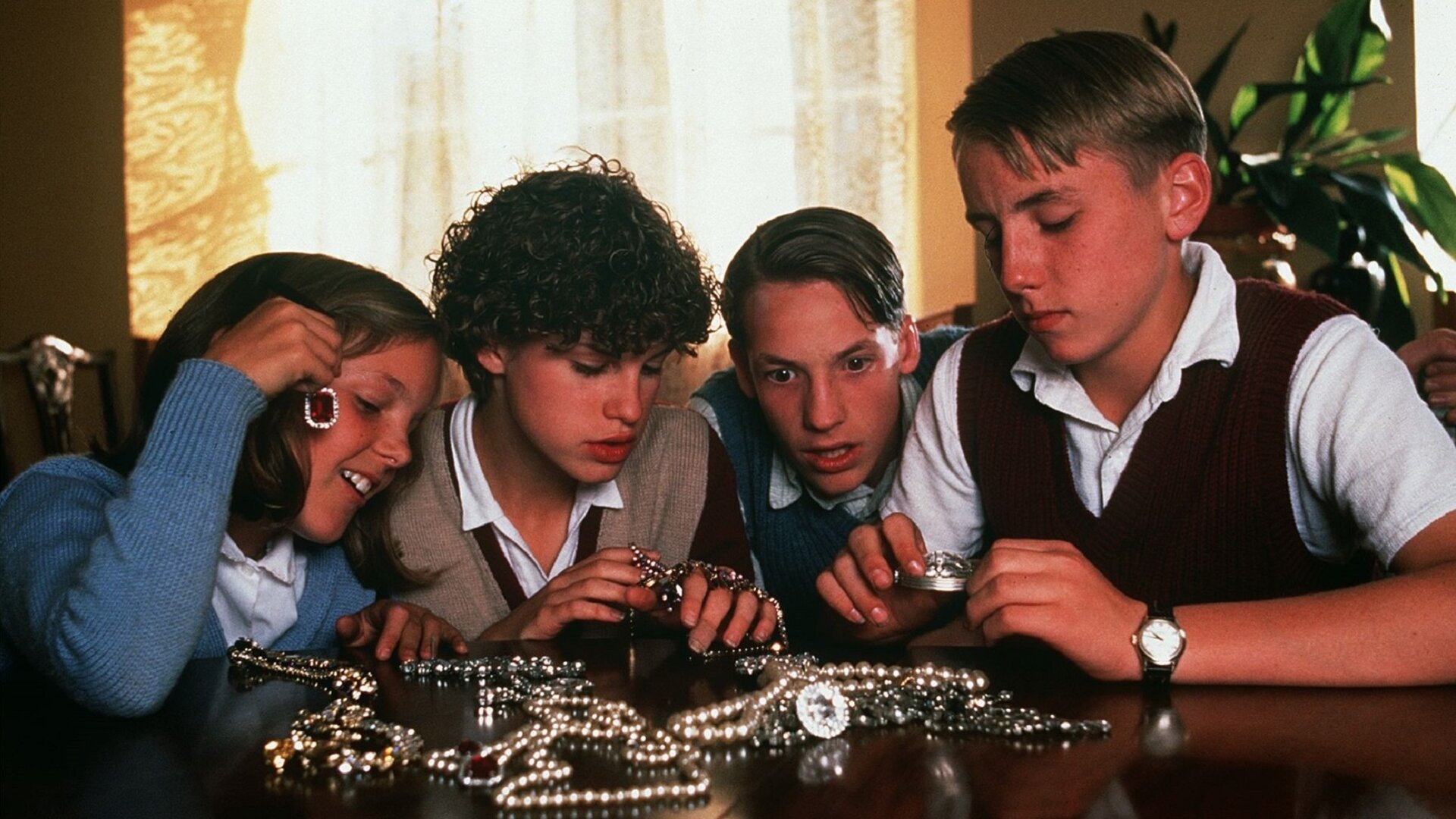 "Fünf Freunde auf großer Fahrt": Anne (Laura Petela) , George (Jemima Rooper), Julian (Marco Williamson) und Dick (Paul Child) sitzen gemeinsam an einem Tisch, auf dem einige Perlenketten und anderer Schmuck liegt. Anne hält sich ein rotes mit Glitzersteinen umrahmtes Schmuckstück an die Nähe ihres Ohrs. Hinter ihnen hängt eine Gardine, rechts daneben steht eine grüne Pflanze.