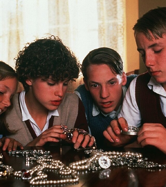 "Fünf Freunde auf großer Fahrt": Anne (Laura Petela) , George (Jemima Rooper), Julian (Marco Williamson) und Dick (Paul Child) sitzen gemeinsam an einem Tisch, auf dem einige Perlenketten und anderer Schmuck liegt. Anne hält sich ein rotes mit Glitzersteinen umrahmtes Schmuckstück an die Nähe ihres Ohrs. Hinter ihnen hängt eine Gardine, rechts daneben steht eine grüne Pflanze.