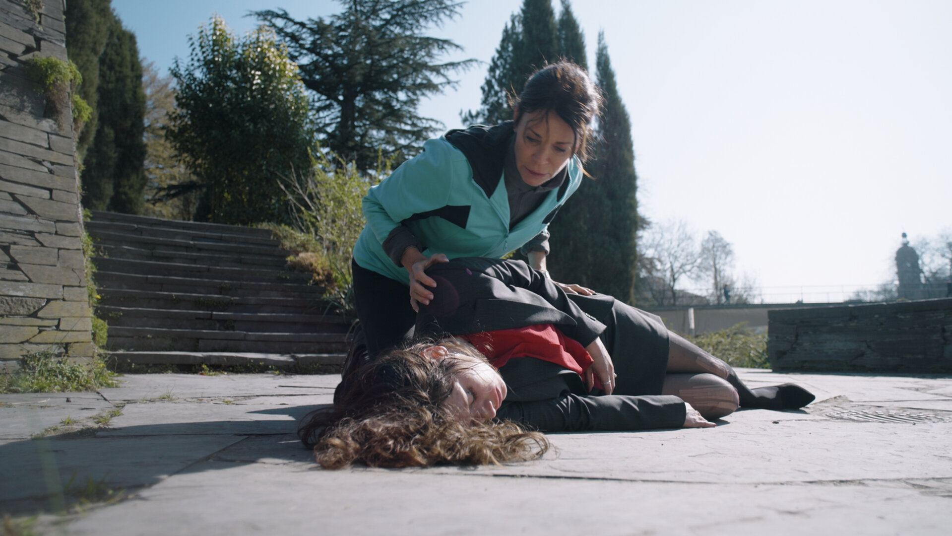 "Notruf Hafenkante - Böses Erwachen": Jasmin Jonas (Gerit Kling) kniet auf dem Boden in einem Park. Sie trägt Sportkleidung. Sie hat sich über eine Frau (Noëmi Krausz) gebeugt, die bewusstlos auf dem Boden liegt.