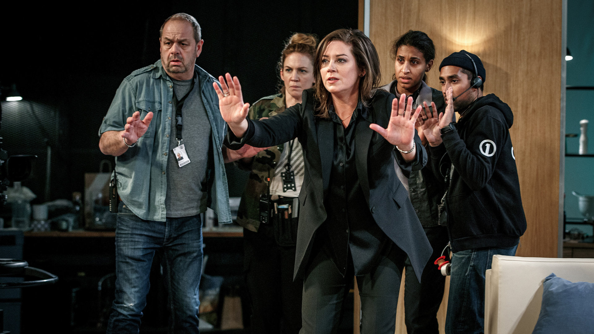 "Kommissar Beck - 58 Minuten": Alex Beijer (Jennie Silfverhjelm) steht mit erhobenen Händen in einem Fernsehstudio. Mimmi (Julia Marko-Nord) und Maya (Lola Zackow) und weitere Kollegen stehen mit angsterfülltem Blick und ebenfalls erhobenen Händen hinter Alex.