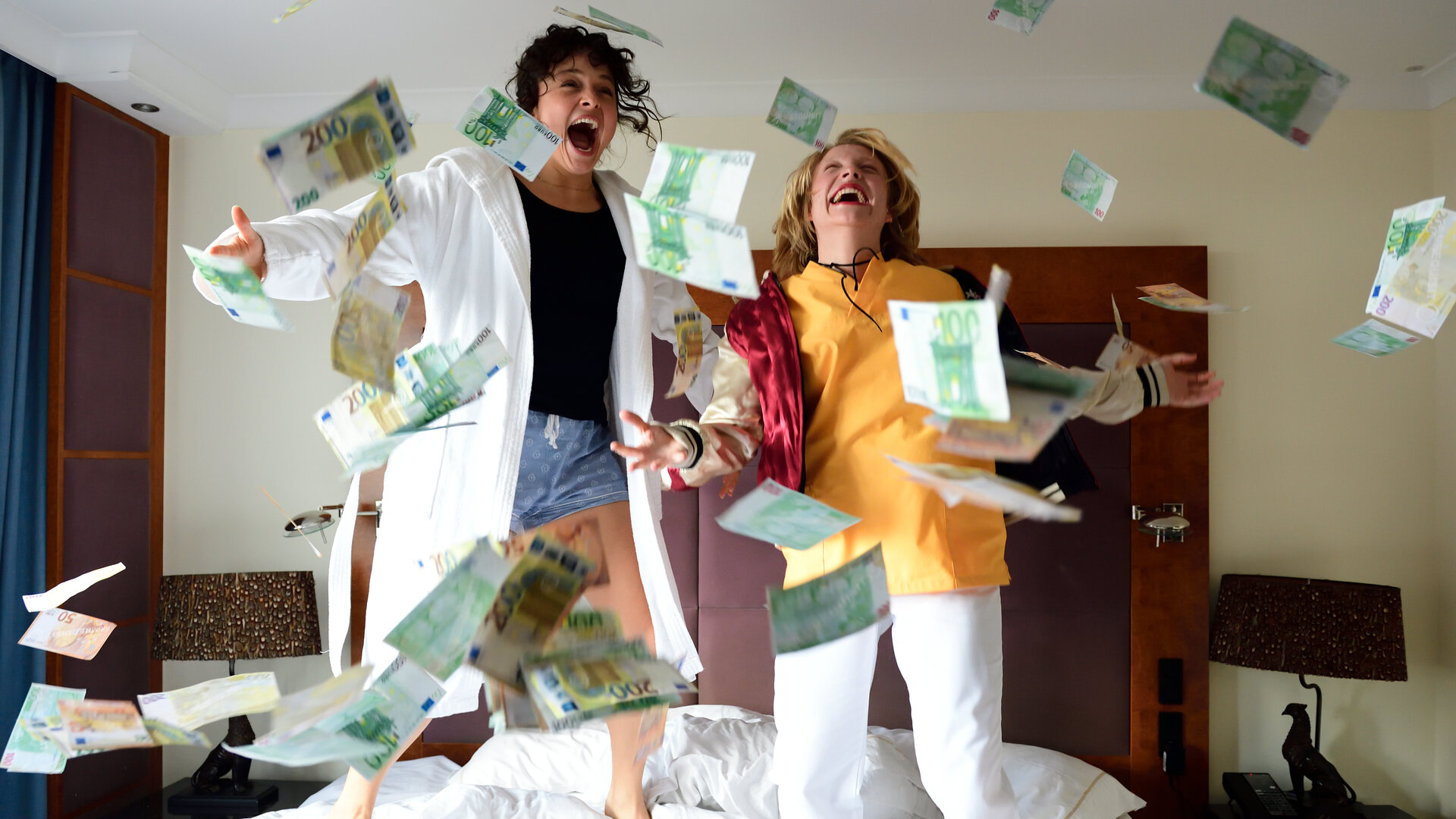 "High Stakes - Dark matter": Ayla (Via Jikeli) und Britta (Pauline Großmann) hüpfen auf dem Bett im Hotelzimmer. Um sie herum wirbeln Geldscheine durch die Luft.