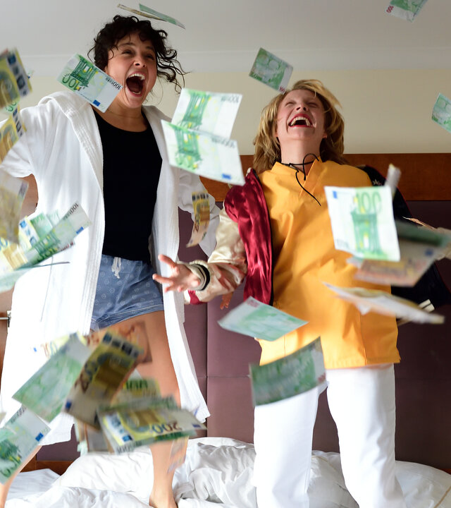 "High Stakes - Dark matter": Ayla (Via Jikeli) und Britta (Pauline Großmann) hüpfen auf dem Bett im Hotelzimmer. Um sie herum wirbeln Geldscheine durch die Luft.