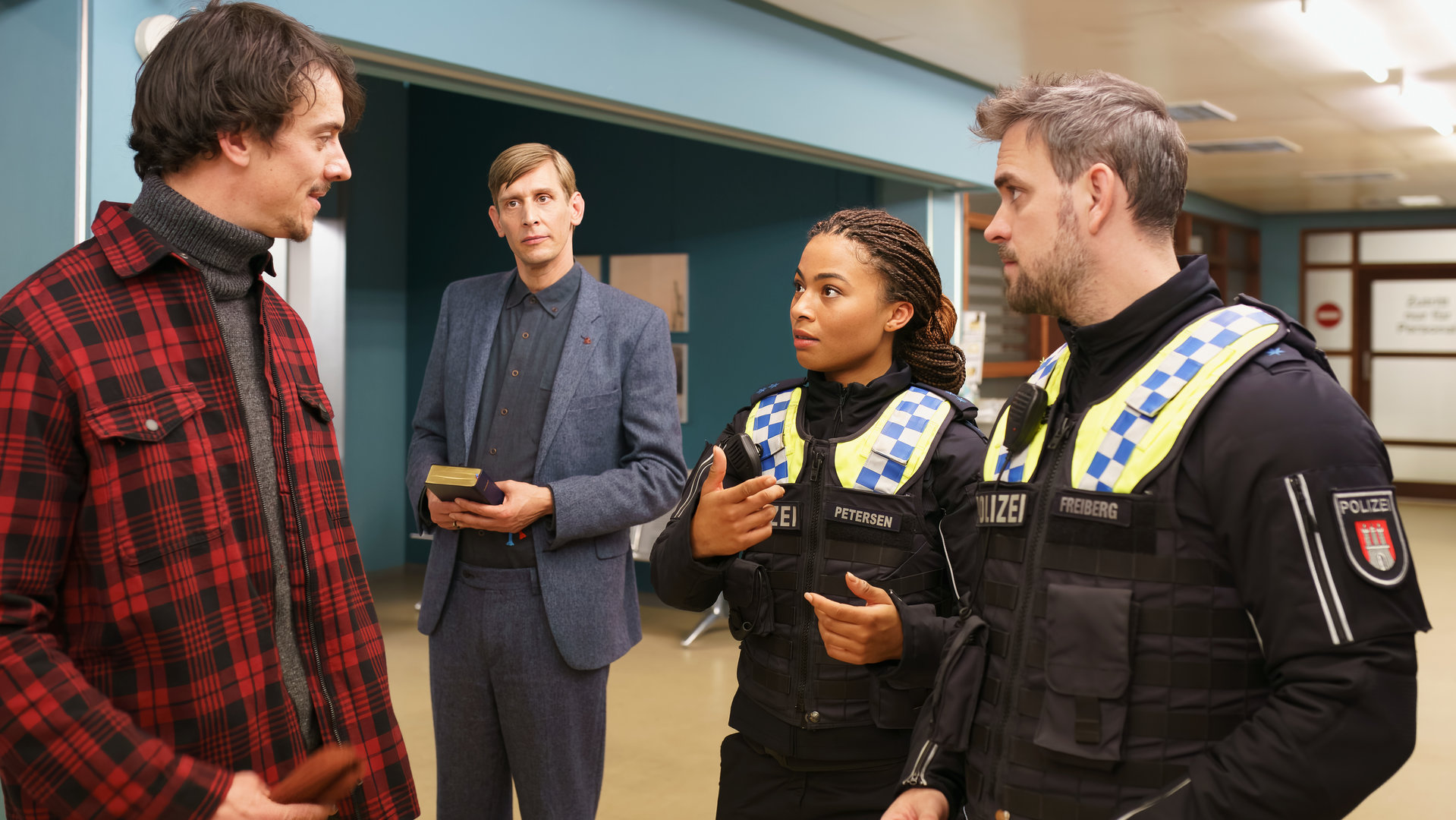 "Notruf Hafenkante - Nächstenliebe": Die Polizistin Daisy Petersen (Aysha Joy Samuel) und ihr Kollege Kris Freiberg (Marc Barthel) stehen nebeneinander auf dem Flur des Elbkrankenhauses. Sie blicken zu Olaf Wemmer (Florian Maria Sumerauer), der ihnen gegenübersteht. Seitlich neben ihnen steht Sven Riemann (Martin Glade). Er hält eine Bibel in den Händen und blickt ebenso zu Olaf Wemmer.