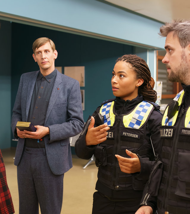 "Notruf Hafenkante - Nächstenliebe": Die Polizistin Daisy Petersen (Aysha Joy Samuel) und ihr Kollege Kris Freiberg (Marc Barthel) stehen nebeneinander auf dem Flur des Elbkrankenhauses. Sie blicken zu Olaf Wemmer (Florian Maria Sumerauer), der ihnen gegenübersteht. Seitlich neben ihnen steht Sven Riemann (Martin Glade). Er hält eine Bibel in den Händen und blickt ebenso zu Olaf Wemmer.