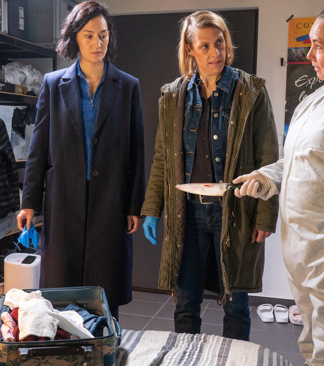 "SOKO Potsdam - Au-Pair": Tamara Meurer (Anja Pahl) und Pauline Hobrecht (Agnes Decker) stehen zusammen mit Spusi Jutta (Gabriele Völsch) im Zimmer des Au-Pairs am Tatort. Jutta hält ein scharfes Messer in der Hand, an dem Blutspuren zu erkennen sind. Das Messer wurde im Koffer des Au-Pairs gefunden, der vor ihnen auf dem Bett liegt.
