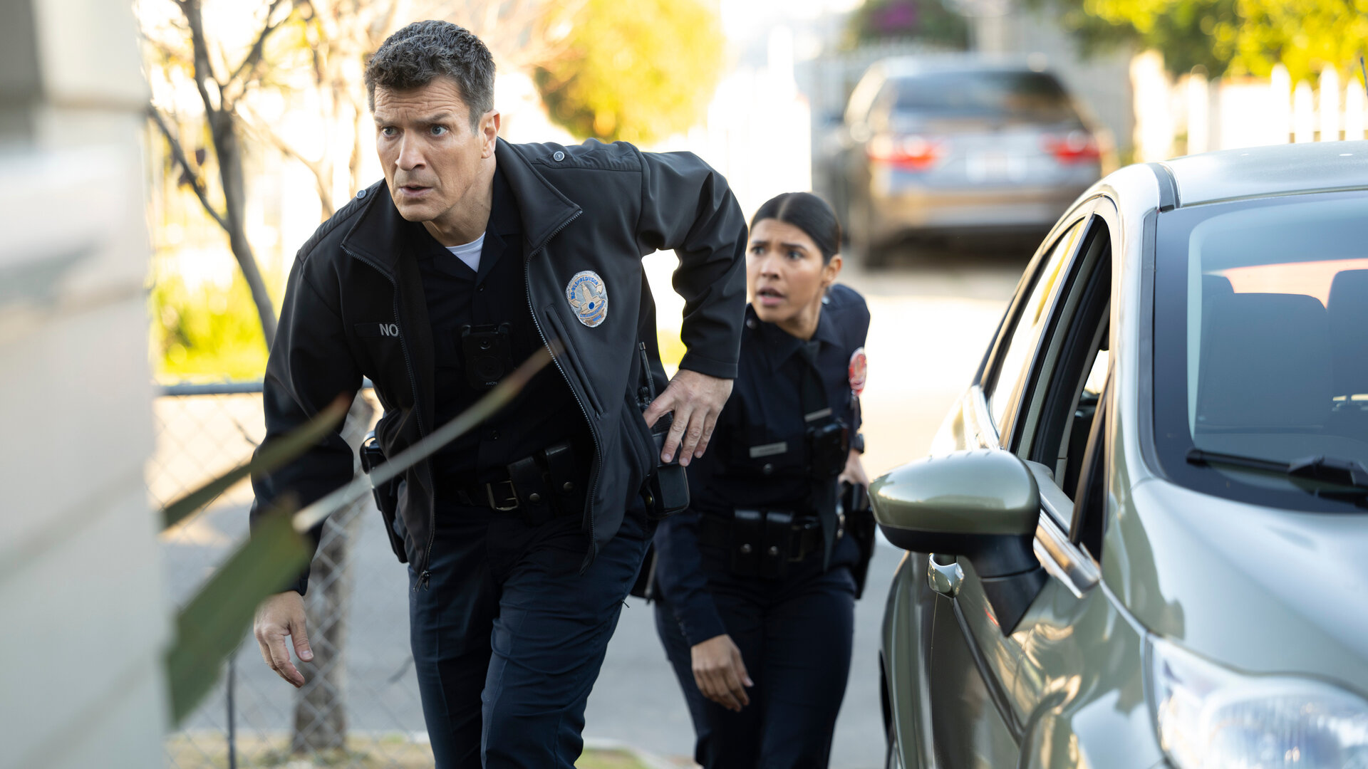 "The Rookie - Geheimnisse und Lügen": Officer Juarez (Lisseth Chavez) und Nolan (Nathan Fillion) schleichen sich auf ein Privatgrundstück und blicken konzentriert in dieselbe Richtung, während jeder von ihnen seine Hand an der Dienstwaffe hat.
