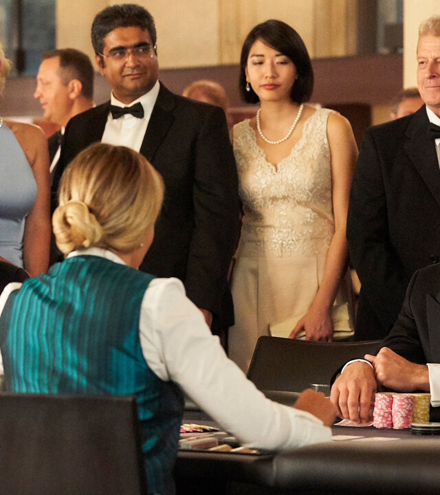 "Suits - Walfang": Harvey (Gabriel Macht, r.) sitzt in einem eleganten Anzug an einem Spieltisch und lächelt die vor ihm sitzende Croupière an. In einer Hand hält er einige Jetons. Hinter ihm stehen Personen in festlicher Abendgarderobe. Links neben ihm sitzt der etwas frustriert dreinschauende Louis (Rick Hoffmann).