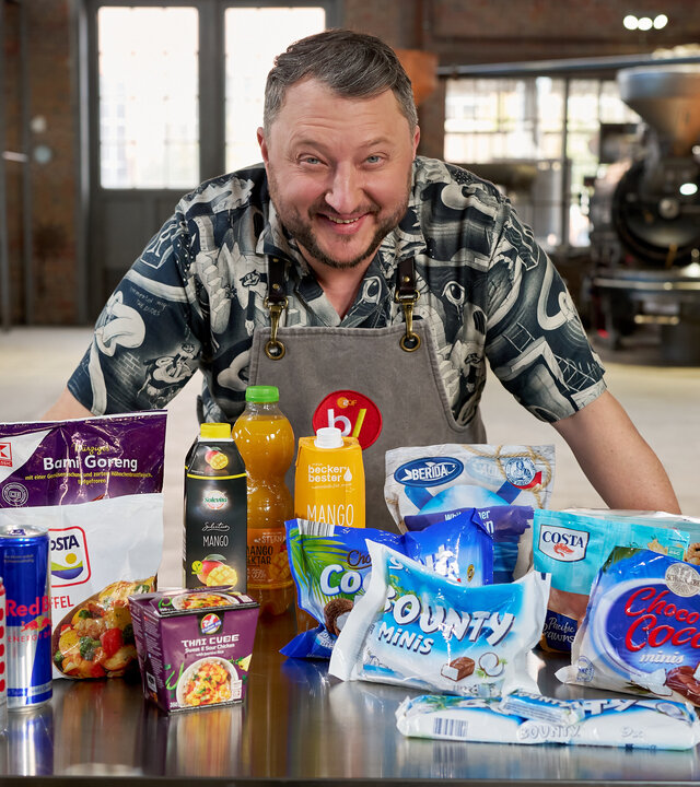 "besseresser: Tricks der Lebensmittelindustrie weltweit - Red Bull, Bounty & Co.": Sebastian Lege steht in seiner Werkstatt, vor ihm auf einem Tisch stehen diverse Lebensmittel.