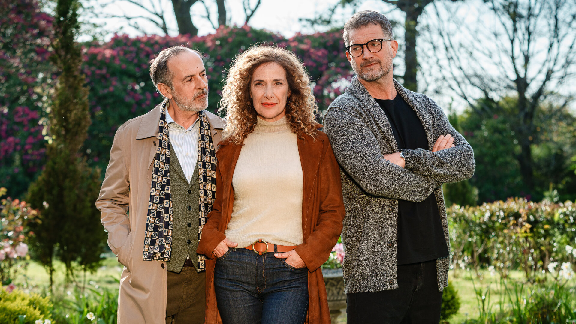 "Rosamunde Pilcher: Wo Liebe aufhört und wo sie beginnt": In einem Garten mit blühenden Pflanzen: Kate Evans (Clelia Sarto) steht in der Mitte zwischen Andrew Evans (Simon Böer) und Yamen Nicholls (Merab Ninidze). Kate hat die Hände in den Hosentaschen und blickt lächelnd in die Kamera. Andrew hat die Arme vor der Brust verschränkt, den Kopf zur Seite gedreht, so dass er Kate anschauen kann. Yamen Nicholls (Merab Ninidze) blickt Kate erwartungsvoll an, während er seine Hände in den Manteltaschen hat.