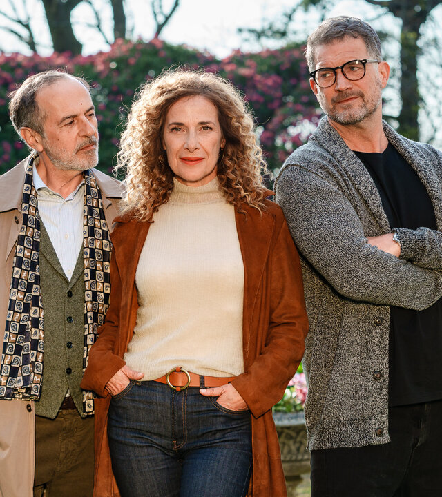 "Rosamunde Pilcher: Wo Liebe aufhört und wo sie beginnt": In einem Garten mit blühenden Pflanzen: Kate Evans (Clelia Sarto) steht in der Mitte zwischen Andrew Evans (Simon Böer) und Yamen Nicholls (Merab Ninidze). Kate hat die Hände in den Hosentaschen und blickt lächelnd in die Kamera. Andrew hat die Arme vor der Brust verschränkt, den Kopf zur Seite gedreht, so dass er Kate anschauen kann. Yamen Nicholls (Merab Ninidze) blickt Kate erwartungsvoll an, während er seine Hände in den Manteltaschen hat.